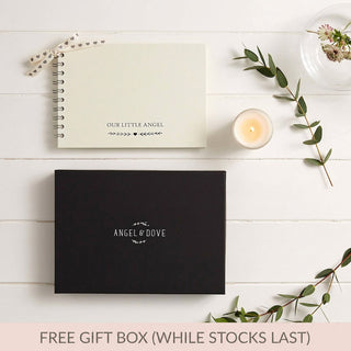 A flat-lay of a gift set on white wood planks, featuring a cream-colored spiral notebook with 'OUR LITTLE ANGEL' printed on it, a black gift box with 'ANGEL & DOVE' in white, a lit candle, stationery cards, and green eucalyptus sprigs. A pink banner at the bottom reads 'FREE GIFT BOX (WHILE STOCKS LAST)'.