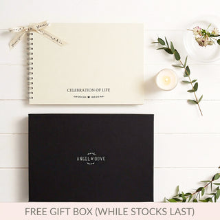 An Angel & Dove celebration of life funeral memory condolence book with a cream spiral-bound cover and a black gift box, styled on a white wood background with a candle, flowers, greenery, and a promotional banner at the bottom.