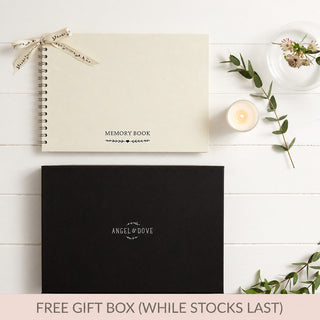 A flat-lay of a memorial keepsake gift set on a white wood background, featuring a cream-colored spiral-bound A4 ivory memory book with a ribbon, a black Angel & Dove branded gift box, a lit candle, and decorative greenery. A pink banner at the bottom reads 'FREE GIFT BOX (WHILE STOCKS LAST)'.