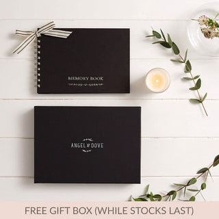 A styled flat-lay of an Angel & Dove A5 black funeral memory book with a striped ribbon, a matching black gift box labelled 'Angel & Dove', a candle, cards, and greenery on a white wood background, with text at the bottom reading 'FREE GIFT BOX (WHILE STOCKS LAST)'.
