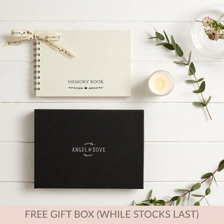 A styled flat-lay of a gift set on a white wood background, featuring a cream spiral-bound memory book tied with a ribbon, a black Angel & Dove gift box, candle, cards, and greenery. A pink banner at the bottom reads 'FREE GIFT BOX (WHILE STOCKS LAST)'.