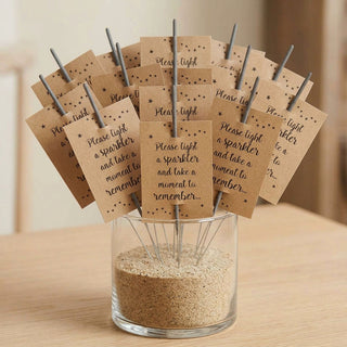 A set of 25 kraft sparkler card funeral favours with star details, slipped onto sparklers and displayed in a clear glass vase filled with sand on a light wood surface.