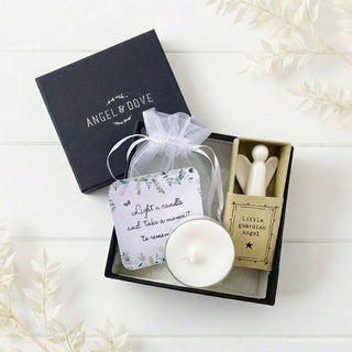 A gift set in an open black box on a white wood background, featuring a black lid printed with 'ANGEL & DOVE,' a white candle, a small floral tag reading 'Light a candle and take a moment to remember,' a small wooden angel figurine in a box labeled 'Little Guardian angel,' and a sheer white drawstring pouch.