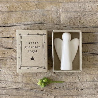 A small white ceramic angel figurine displayed inside an open matchbox-style box next to its lid, which reads 'Little guardian angel' with a star. The items are arranged on a rustic wooden surface with a small green flower stem in front.