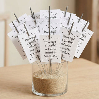 A set of white card funeral sparkler cards with black printed text and star details, slipped through sparklers and arranged in a clear glass jar filled with sand on a light wood surface.